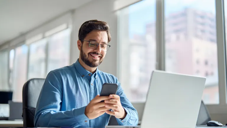 Happy professional business man company employee, smiling busy young businessman working on laptop holding smartphone using cellphone looking at mobile phone sitting at office workplace desk