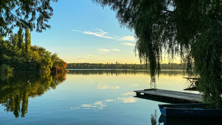 A lake at sundown. On the sides, willows and other trees. A small ship dock is also seen on the right.