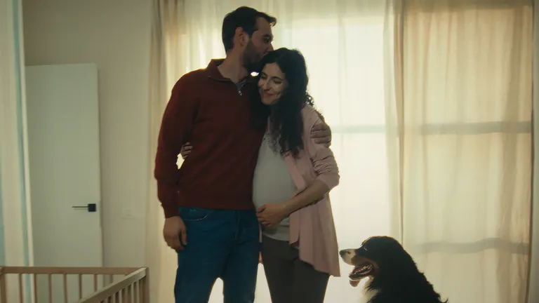 A man kisses a pregnant woman on her head while hugging. They embrace as a family inside a room with a toddler bed while a dog is sitting nearby