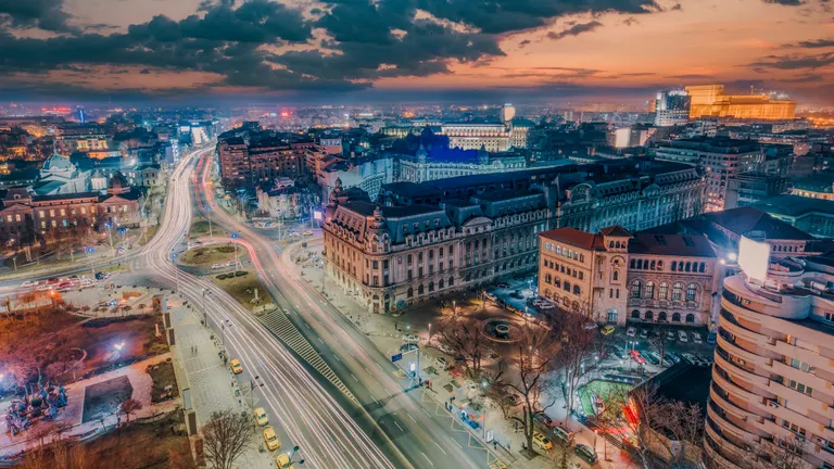 University Square in Bucharest, Capital city of Romania, seen from above. Many building seen, including the Romanian Palace of the Parliament. The sky is orange, at sundown, with scattered clouds