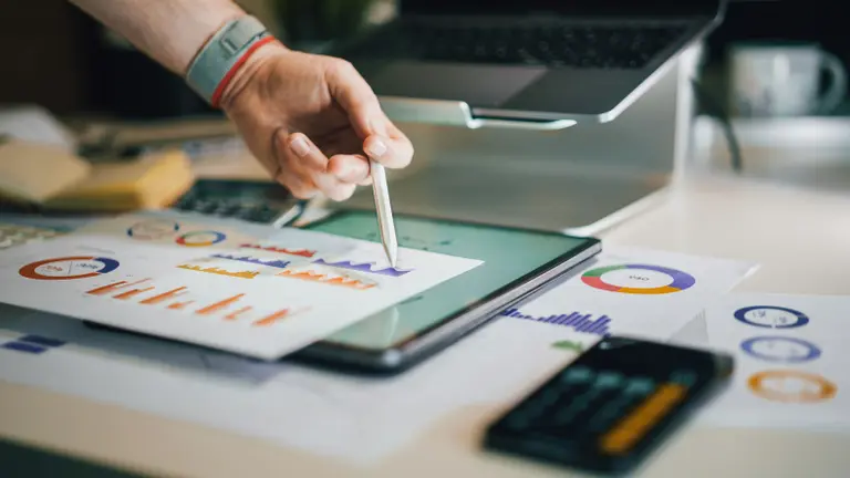 A hand indicates with a pen information on some printed papers containing different colour graphics