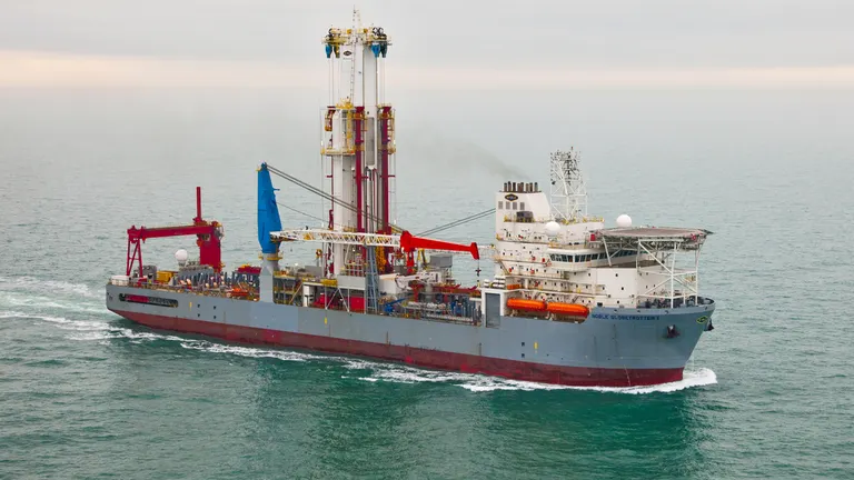 An offshore exploration ship sails in waters. It is gray and red and has a huge installation used to drill exploration and production wells in underwater oil and gas blocks