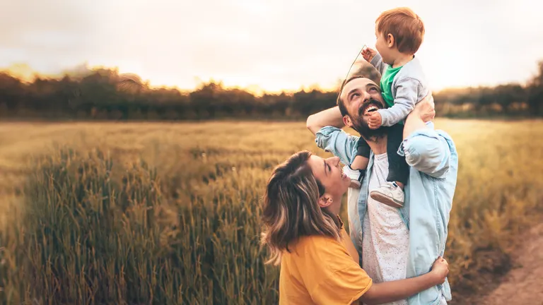 A man with a child on his shoulders and a woman embracing him smile in a wheat field