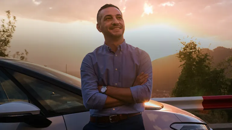 Male OMV Petrom employee next to an electric car with a backdrop of a Romanian landscape during sundown